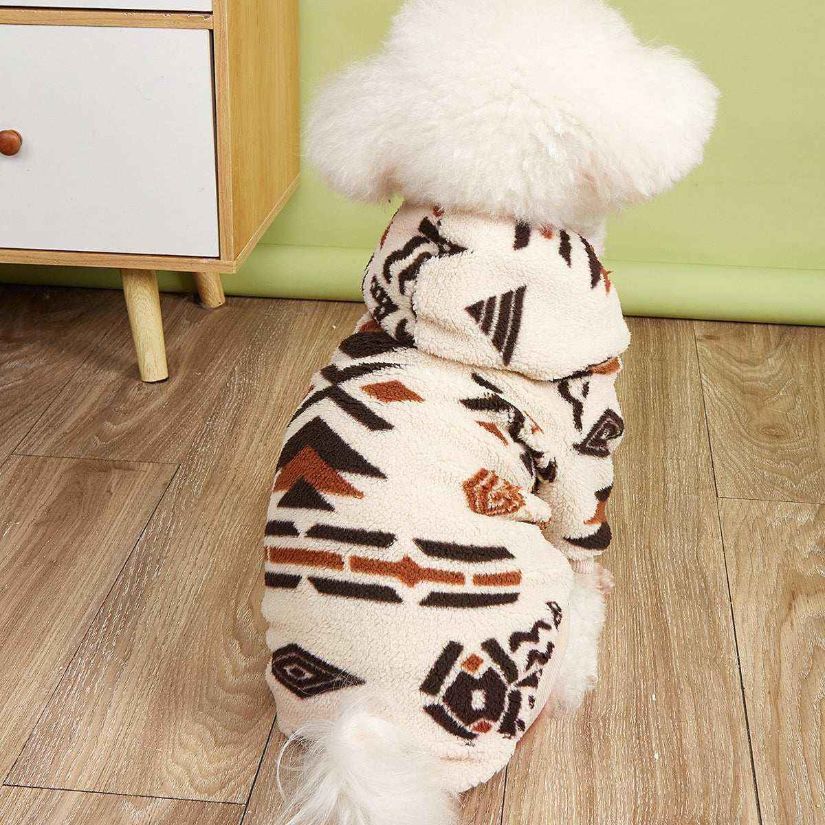 Small white fluffy dog wearing a cozy patterned fleece jacket indoors on wooden floor
