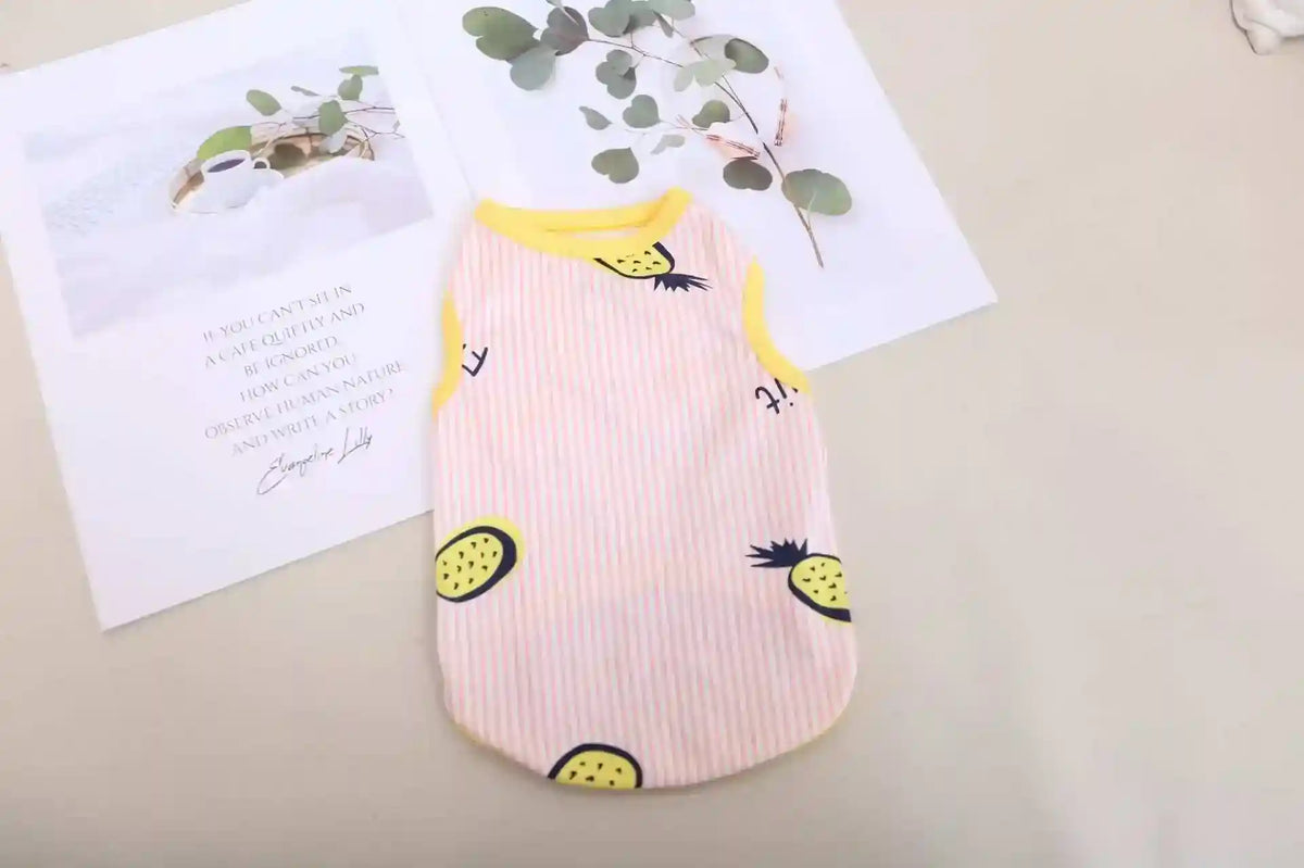 Pink striped sleeveless pet shirt with yellow pineapple print on beige surface with magazine and green leaves