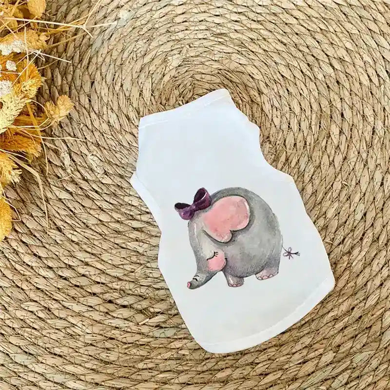 White pet shirt with cute gray elephant wearing purple bow on woven straw mat next to dried flowers