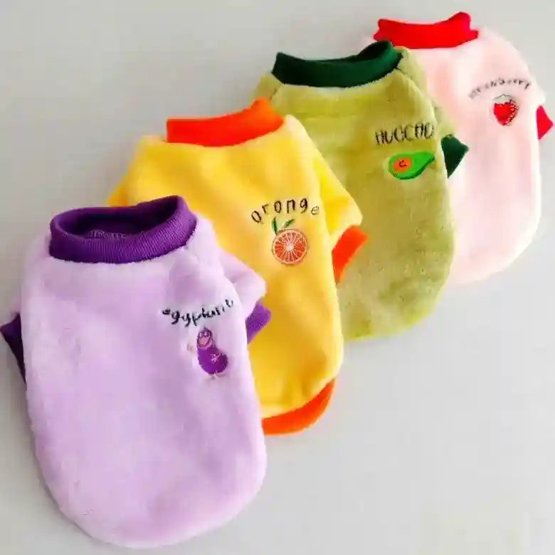 Four colorful plush pet sweaters with fruit and vegetable embroidery on a light surface