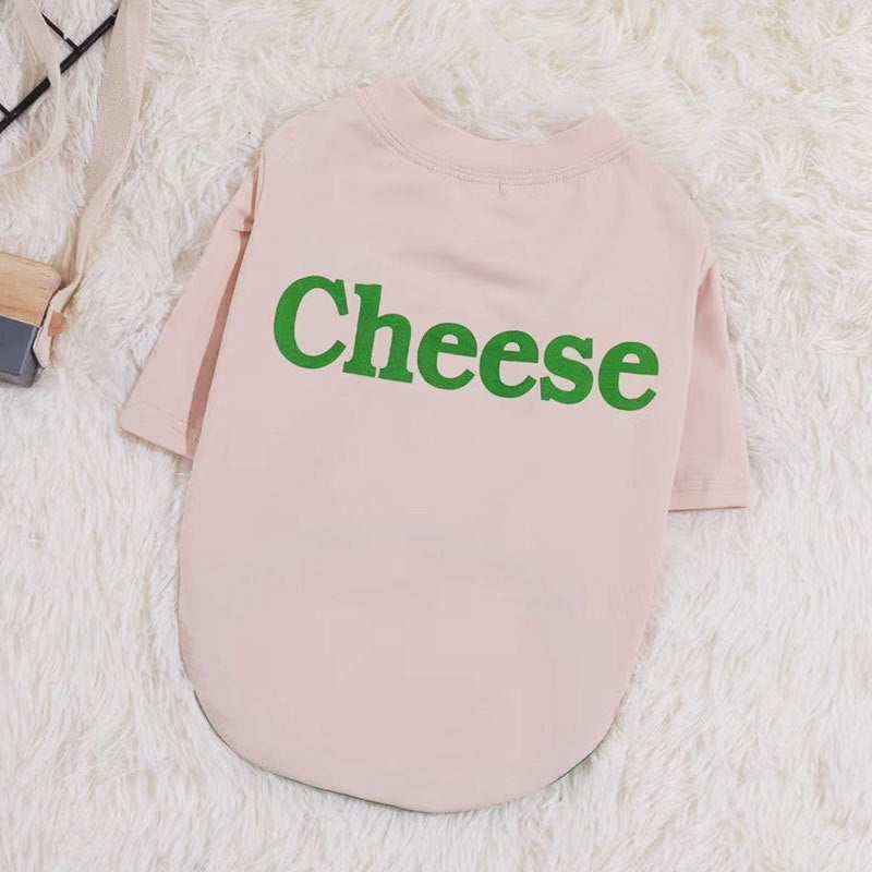 Light pink dog shirt with green text 'Cheese' printed on back, displayed on white fluffy background