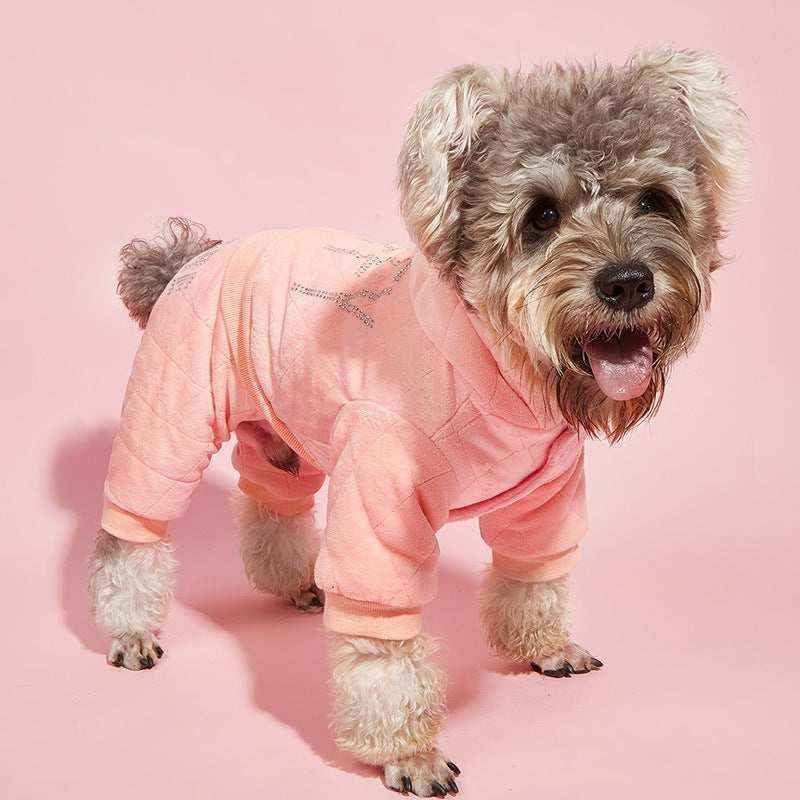 Small fluffy dog wearing a pink cozy jumpsuit standing on a pink background
