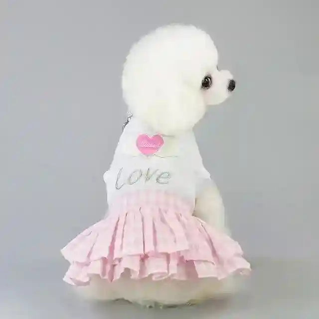 White poodle wearing a white top with 'love' and a pink heart, and a pink checkered skirt dress