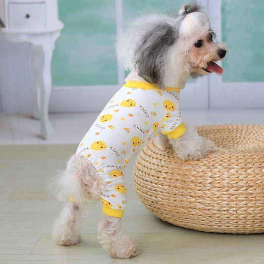 Small dog wearing white and yellow chick-themed pajamas standing with front paws on a woven ottoman indoors