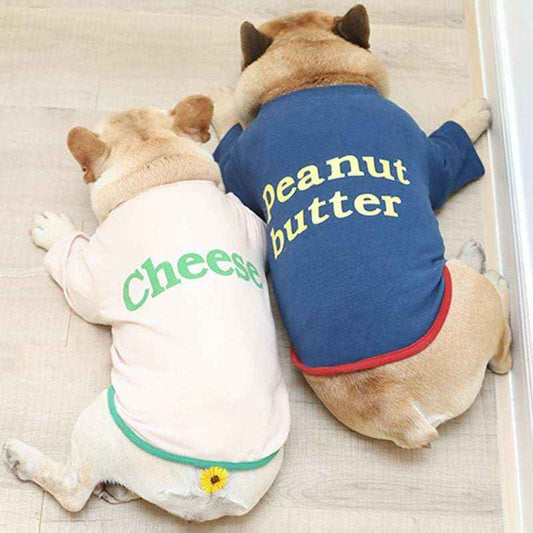 Two small dogs lying on floor, wearing matching shirts labeled 'cheese' and 'peanut butter' in pastel and blue colors.