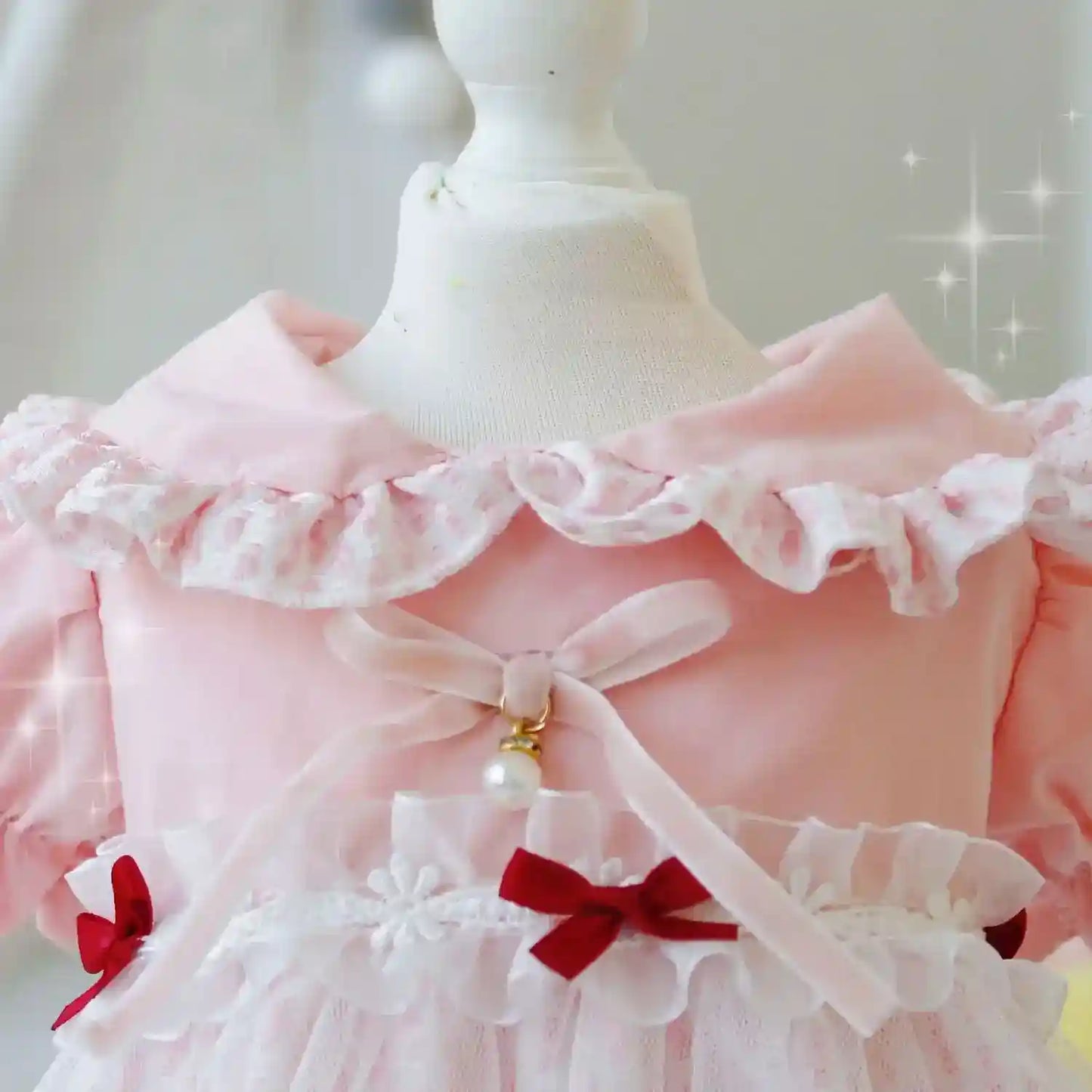 Close-up of pink baby dress with ruffled collar, sheer bow, pearl pendant, and red ribbon accents