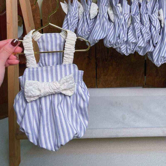 Hand holding striped baby romper with white bow, multiple rompers hanging on rack in background