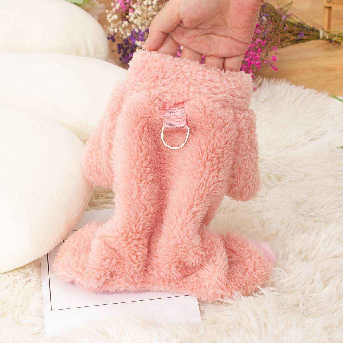 Hand holding a soft pink plush dog harness with metal D-ring on a white furry rug