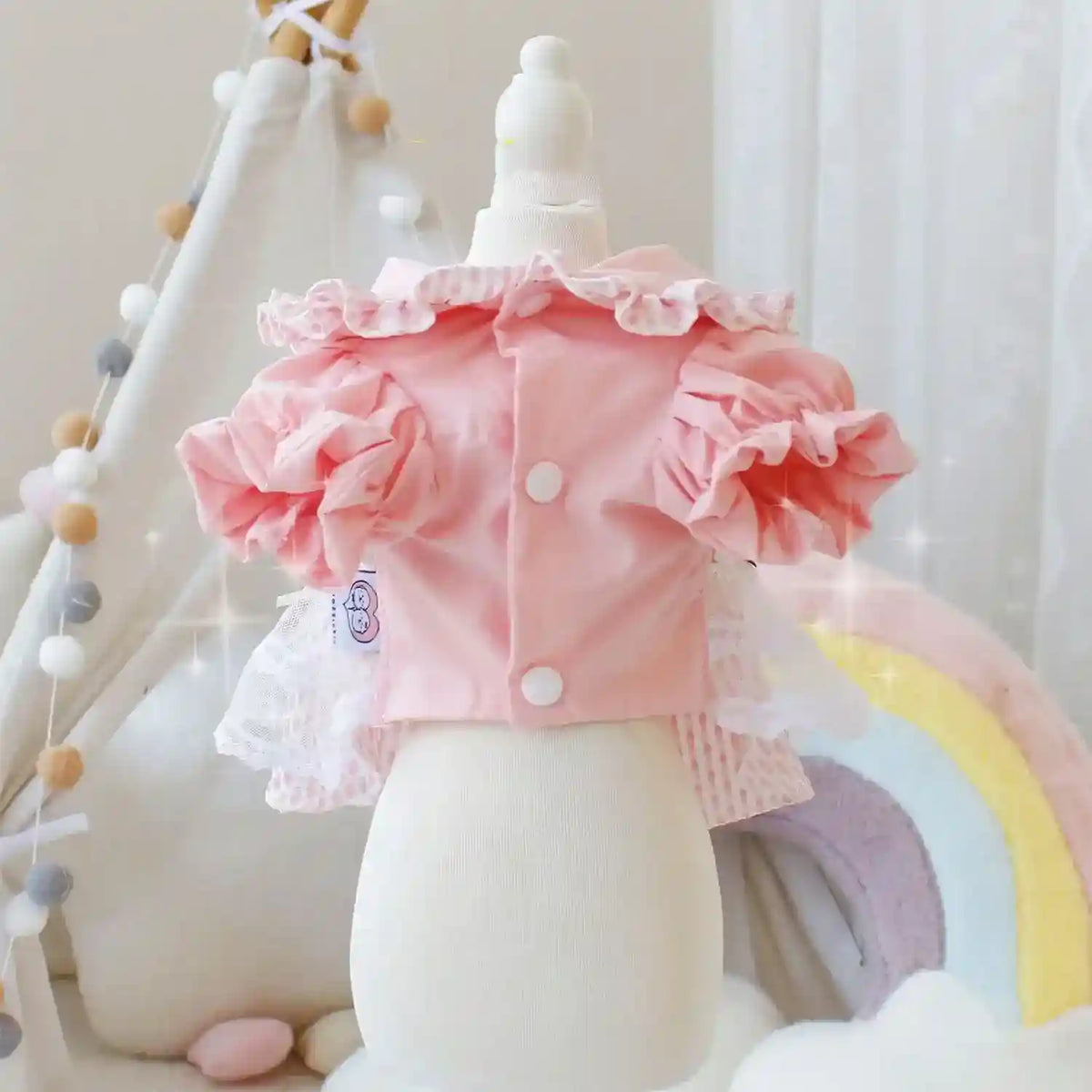 Pink ruffled baby jacket with white buttons on mannequin in pastel nursery with rainbow and teepee