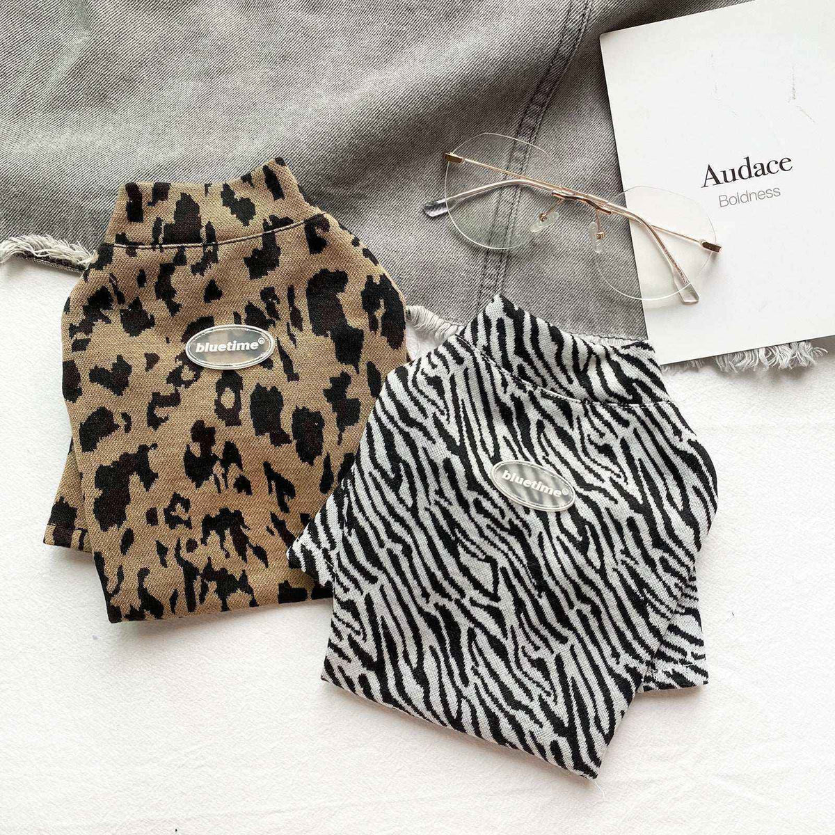 Two folded animal print pet sweaters, leopard and zebra patterns, with bluetime logos, glasses, and gray fabric