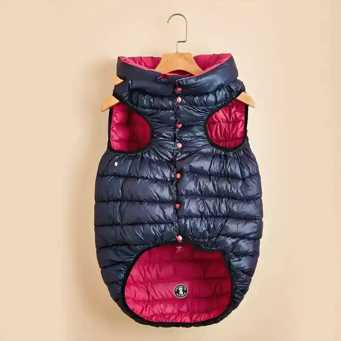 Padded black and pink dog vest with hood hanging on wooden hanger against beige background