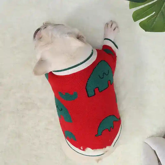 White French Bulldog wearing red sweater with green elephant pattern lying on fluffy white rug
