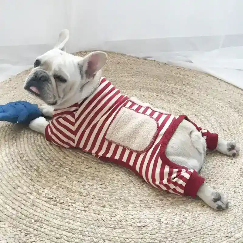 French Bulldog wearing red and white striped dog pajamas lying on woven round rug with blue toy
