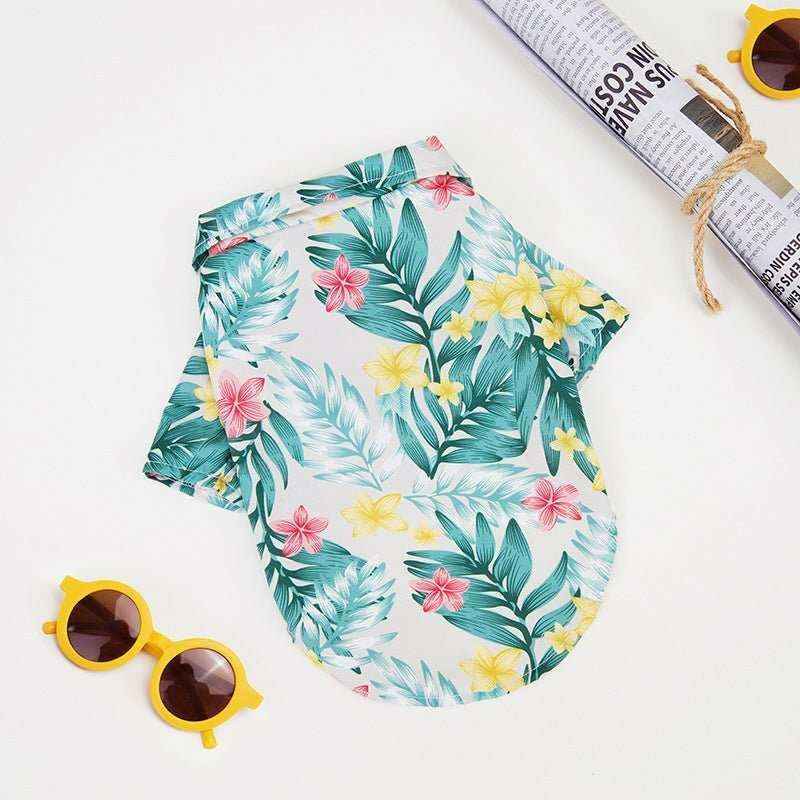 Hawaiian style dog shirt with green leaves and yellow and pink flowers, lying flat with yellow sunglasses and rolled newspaper