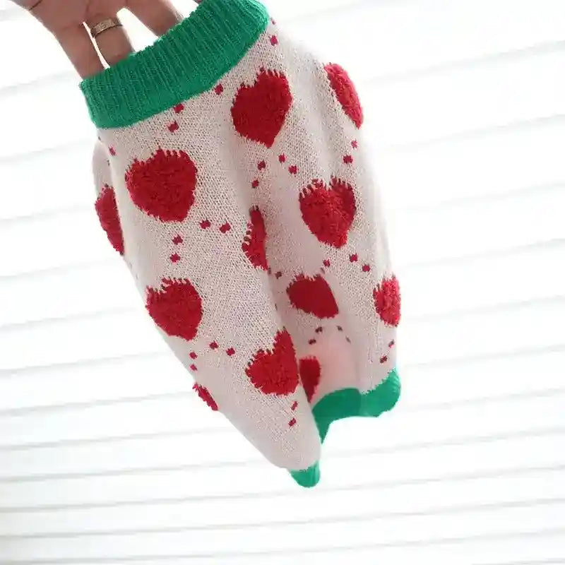 White knitted dog sweater with red heart patterns and green trim, held against light background