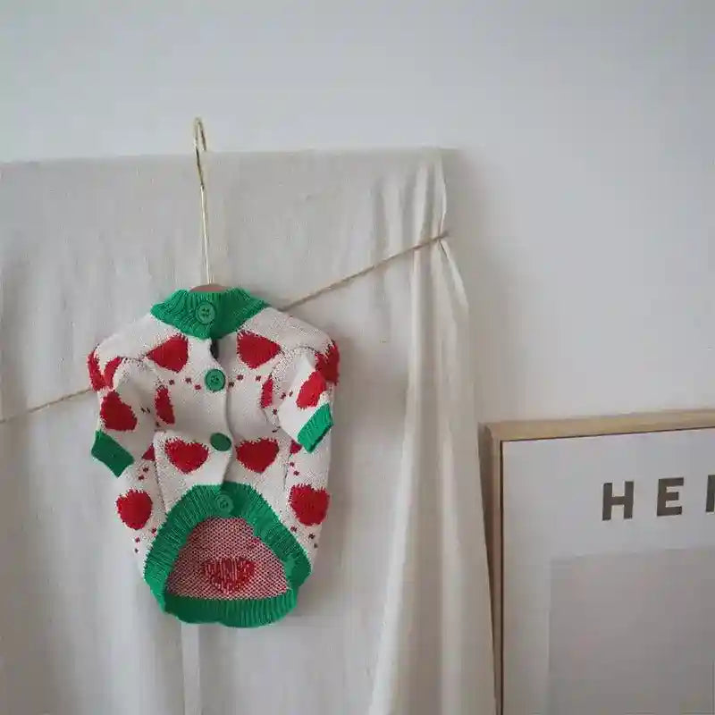 Knitted dog cardigan with red hearts and green trim hanging on a hook against a beige fabric background