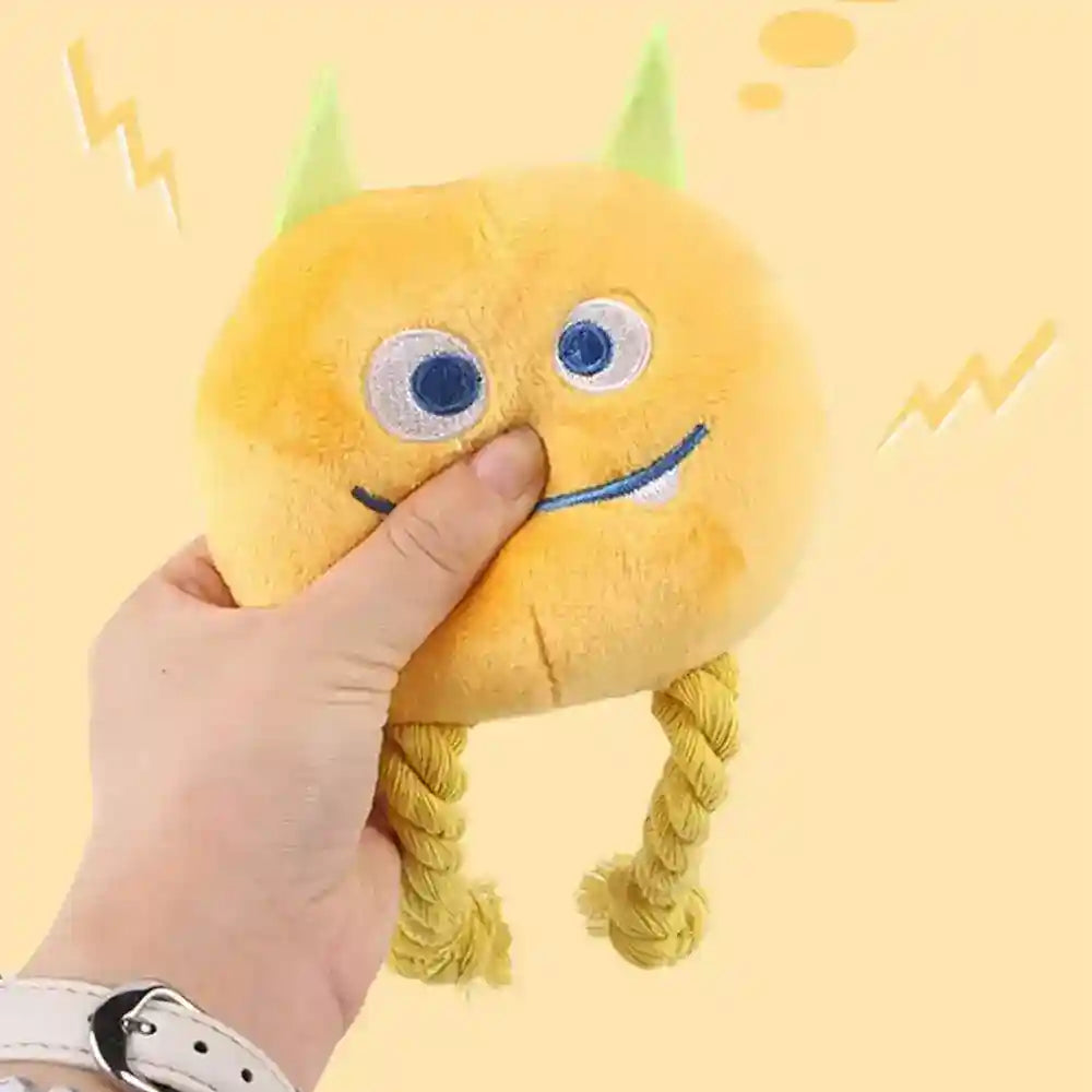 Hand holding yellow plush dog toy with blue eyes, green horns, and rope legs on yellow background