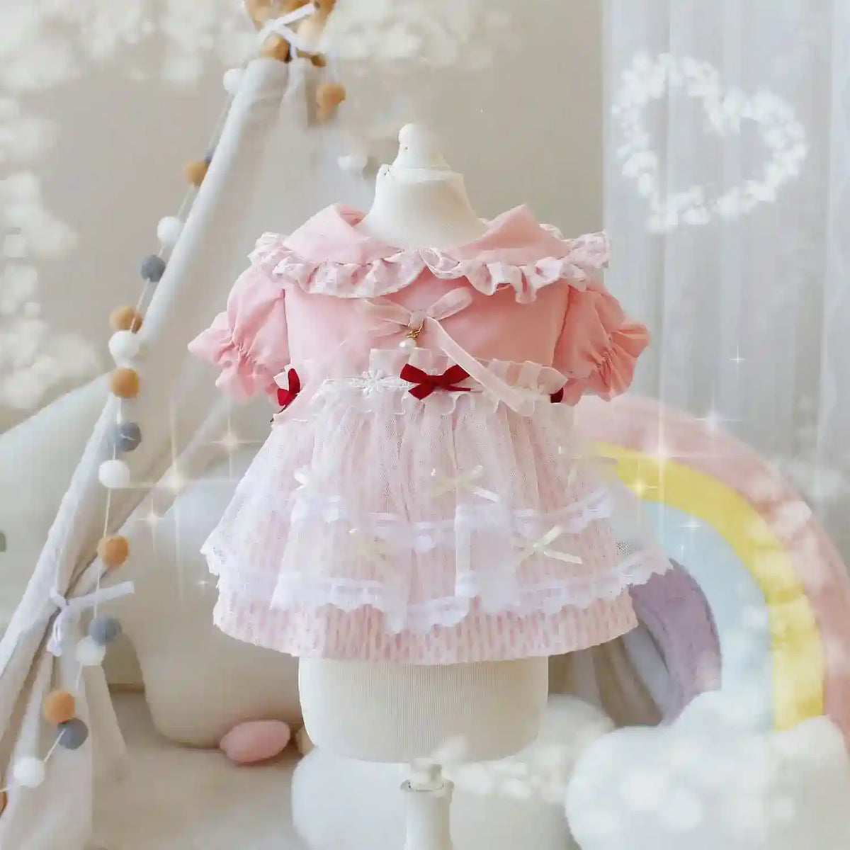Pink baby dress with lace, bows, and ruffled collar on mannequin in whimsical nursery with rainbow and teepee