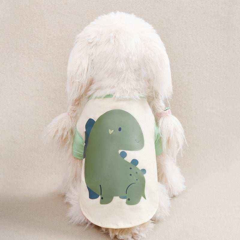 small white fluffy dog wearing a cream-colored shirt with a cute green dinosaur print on the back
