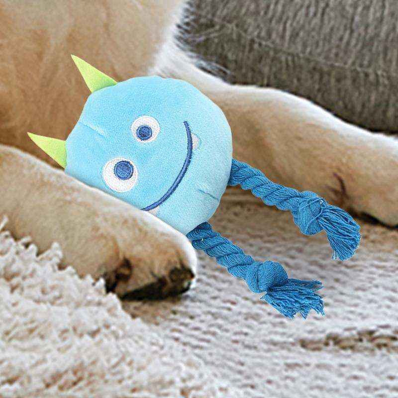 Blue plush dog toy with smiling face and green horns on beige couch near dog's paws