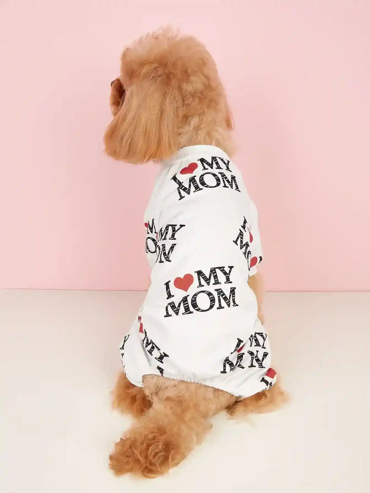 Small curly-haired dog wearing a white pet shirt with I love my mom print sitting against pink background