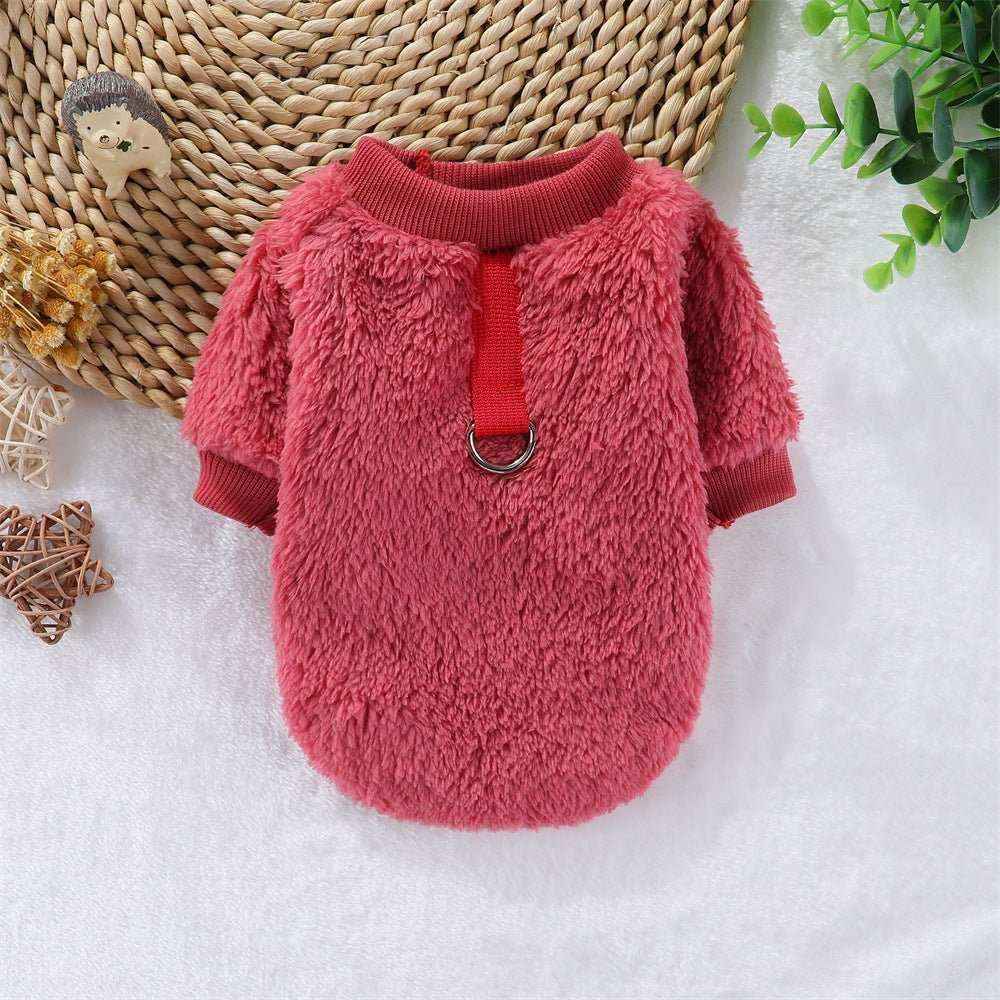 Pink cozy fleece dog sweater with a red collar strap and metal ring on a white surface with decorative wicker and greenery