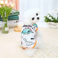 Small white poodle wearing dinosaur print dog shirt sitting on table with flowers and books
